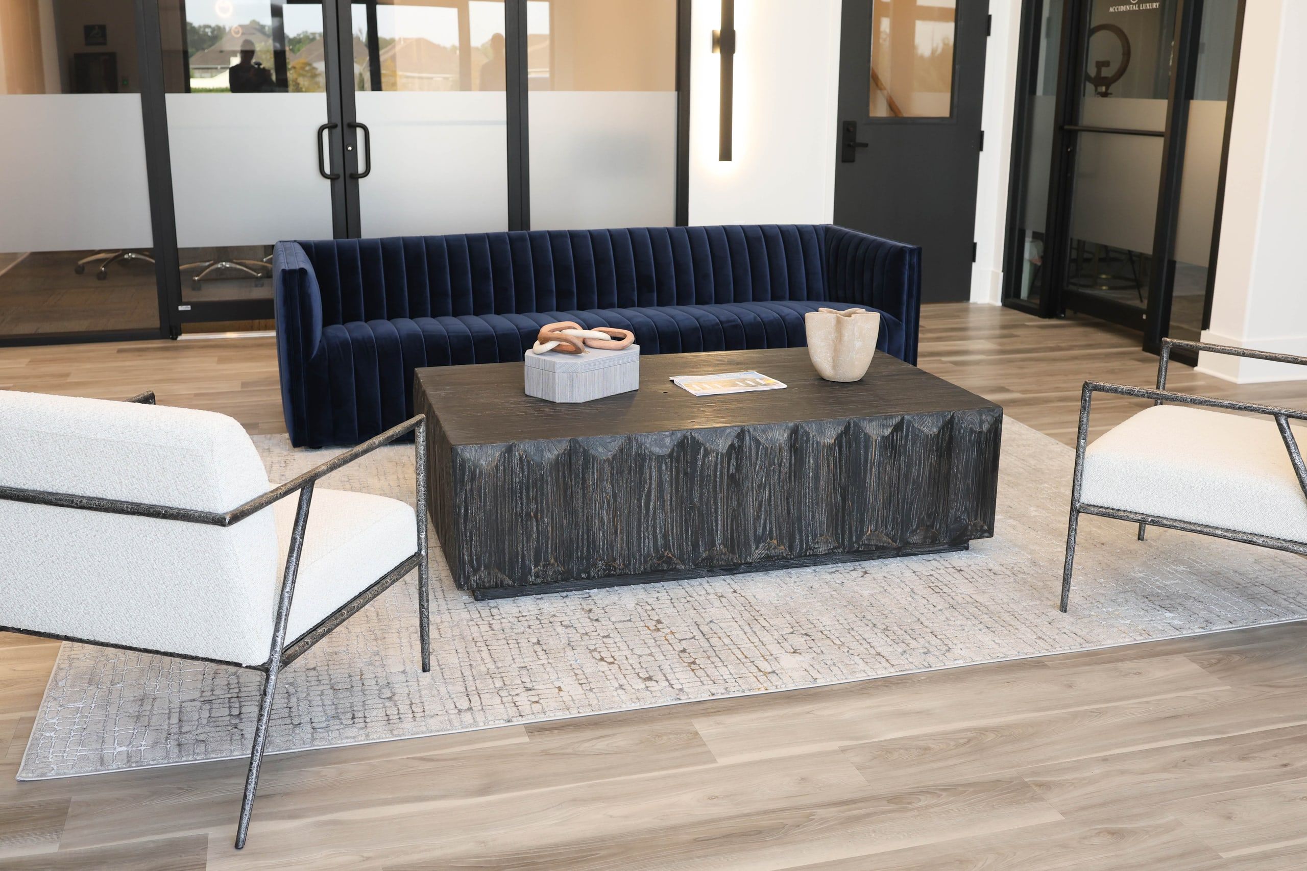 Designer office lobby at The W Office Suites in Prairieville, LA, with a navy velvet sofa, white lounge chairs, and black steel-and-glass suite doors