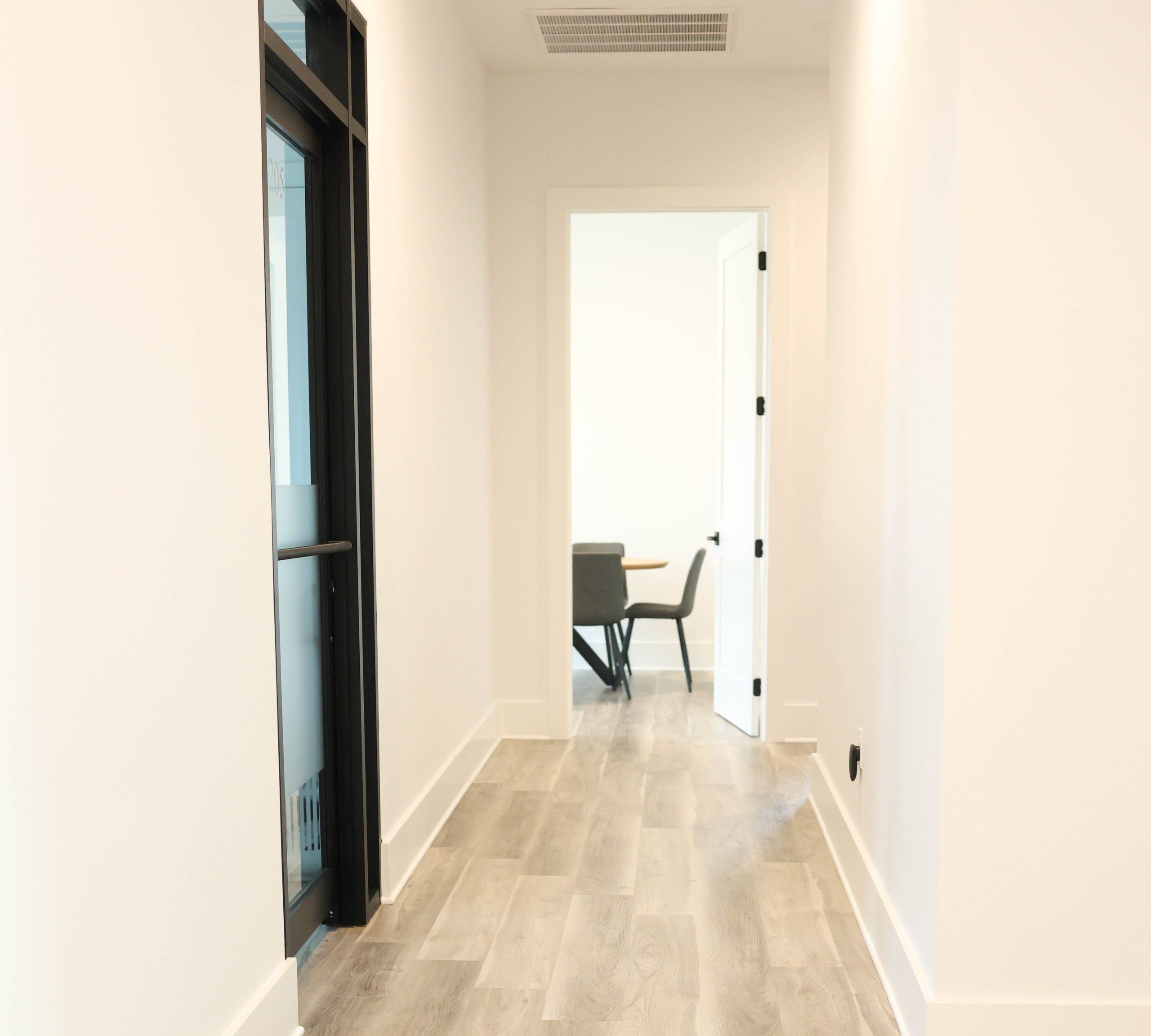 Hallway of private office suites at The W Office Suites, with a black steel-and-glass office door and an open office at the end